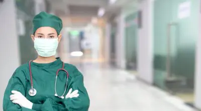 Healthcare worker in scrubs, mask, and gloves standing in a hospital hallway.