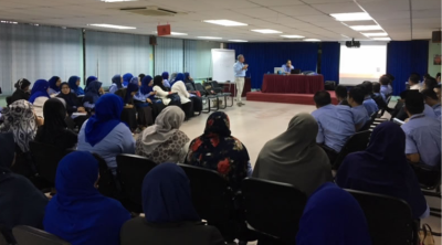 A presenter addresses an audience in a large room with projector screen and whiteboard.