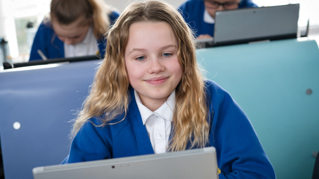 A young student smiles while working on a laptop in a classroom.