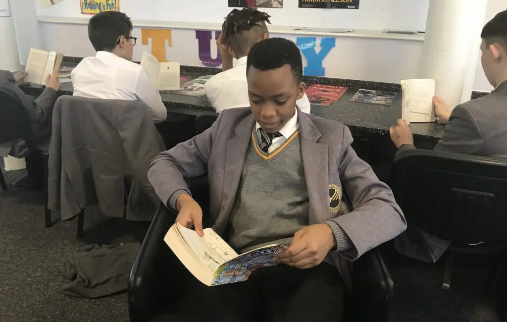 A schoolboy in uniform sits reading a book, surrounded by other students also reading in a classroom setting.