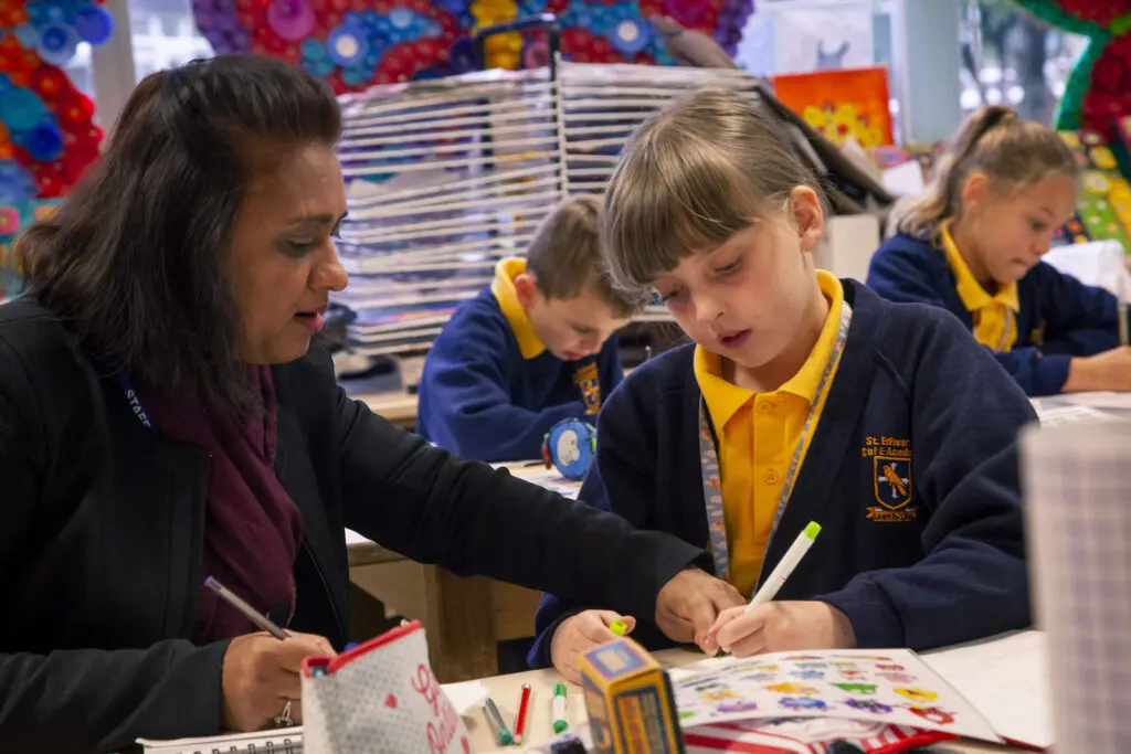 A teacher assists a primary school aged girl with a writing assignment in a classroom.