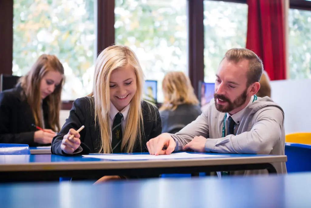 A teacher assisting a smiling student at her desk in a classroom.