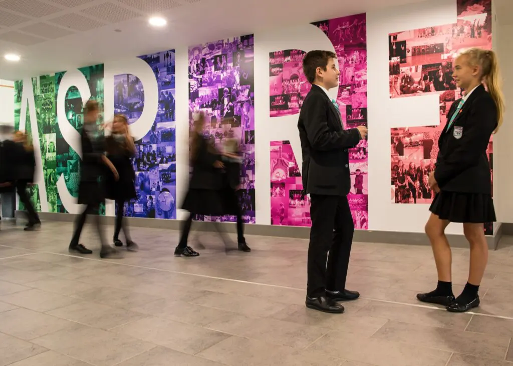Two school students in uniform talking in a hallway with a large, colorful mural on the wall.