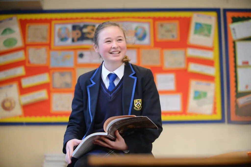 Smiling schoolgirl in uniform holding open textbook in classroom.