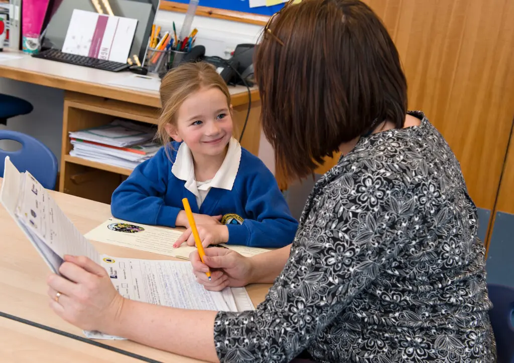 A teacher works with a smiling primary school student at a desk.