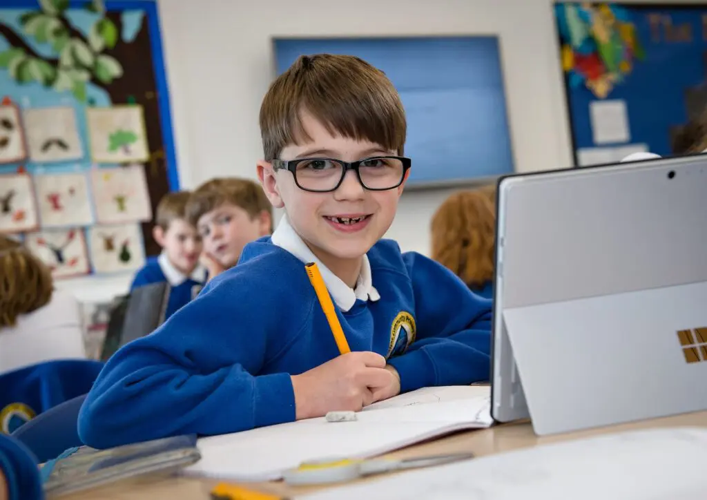 Primary school student in uniform smiles while using a tablet in class.