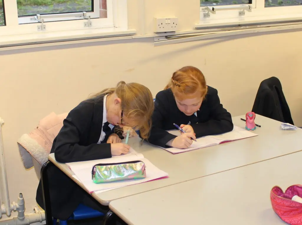 Two schoolgirls in uniform writing in notebooks at a desk in a classroom.