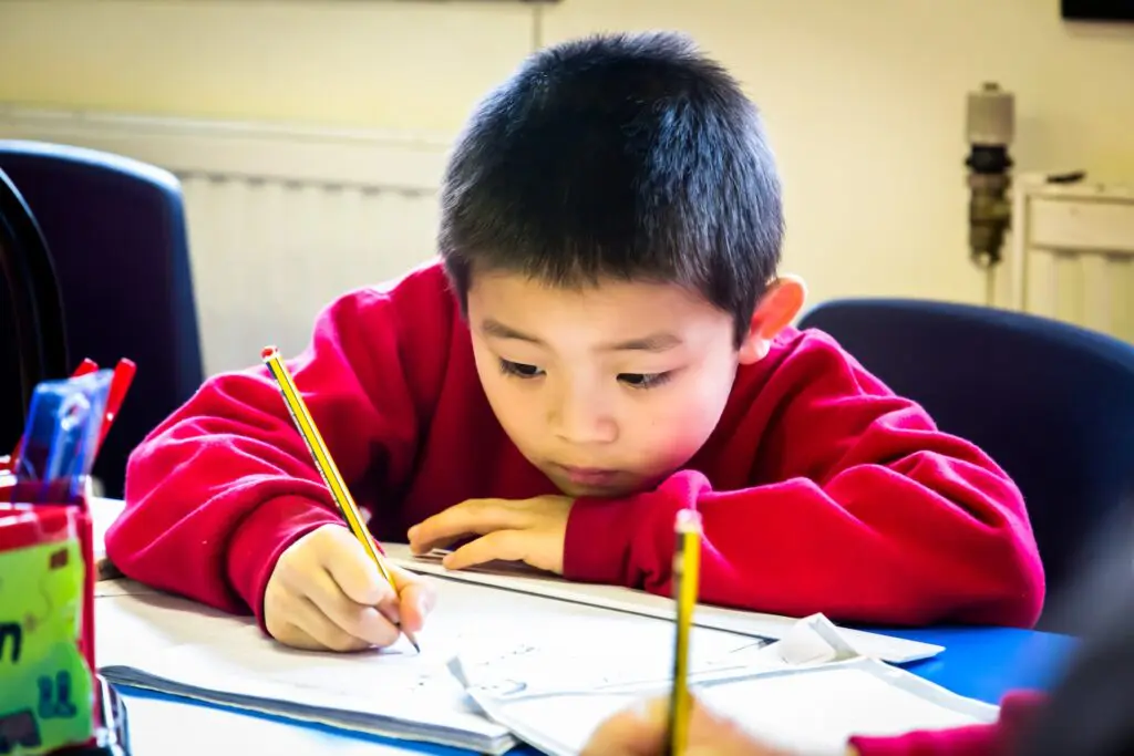 A young boy in a red sweater concentrating while writing with a pencil at his desk.
