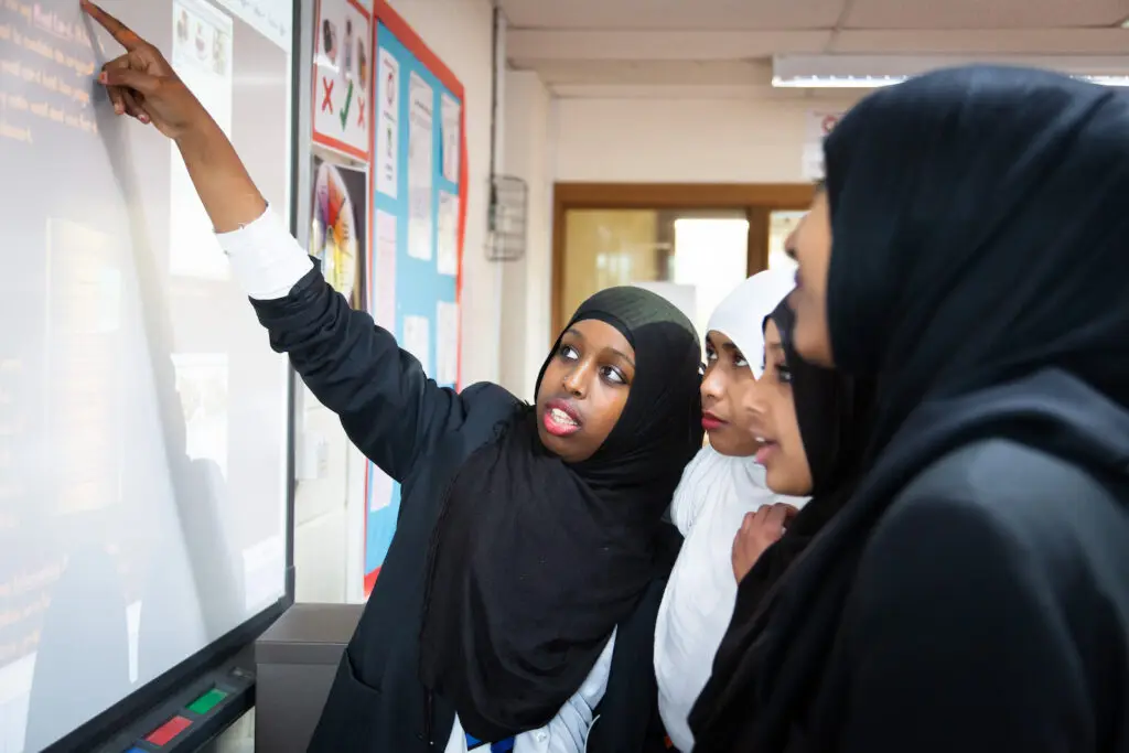 Student presenting at a whiteboard to a group of classmates in a classroom.