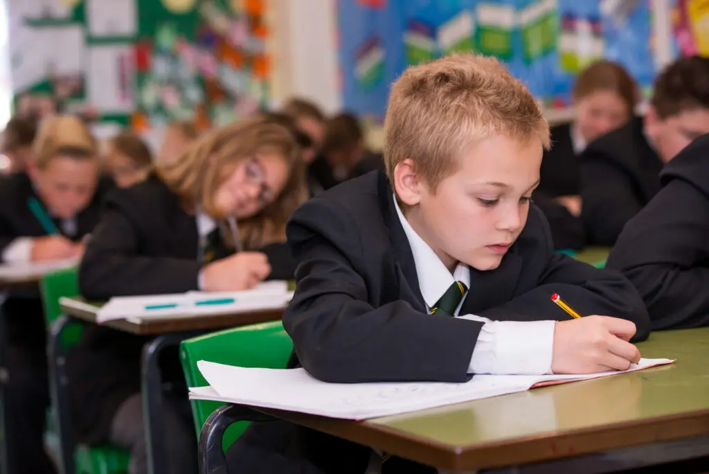 A young student in a school uniform is writing at his desk in a classroom.