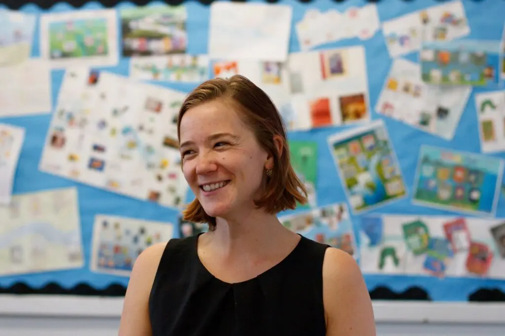 A woman smiling in front of a bulletin board covered in children's artwork.